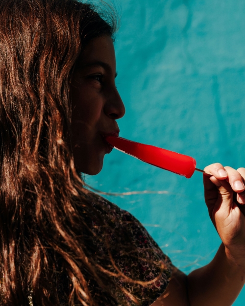 Side view of a young woman with long brown hair enjoying a red popsicle against a bright turquoise wall, highlighting summer treat and casual joy.