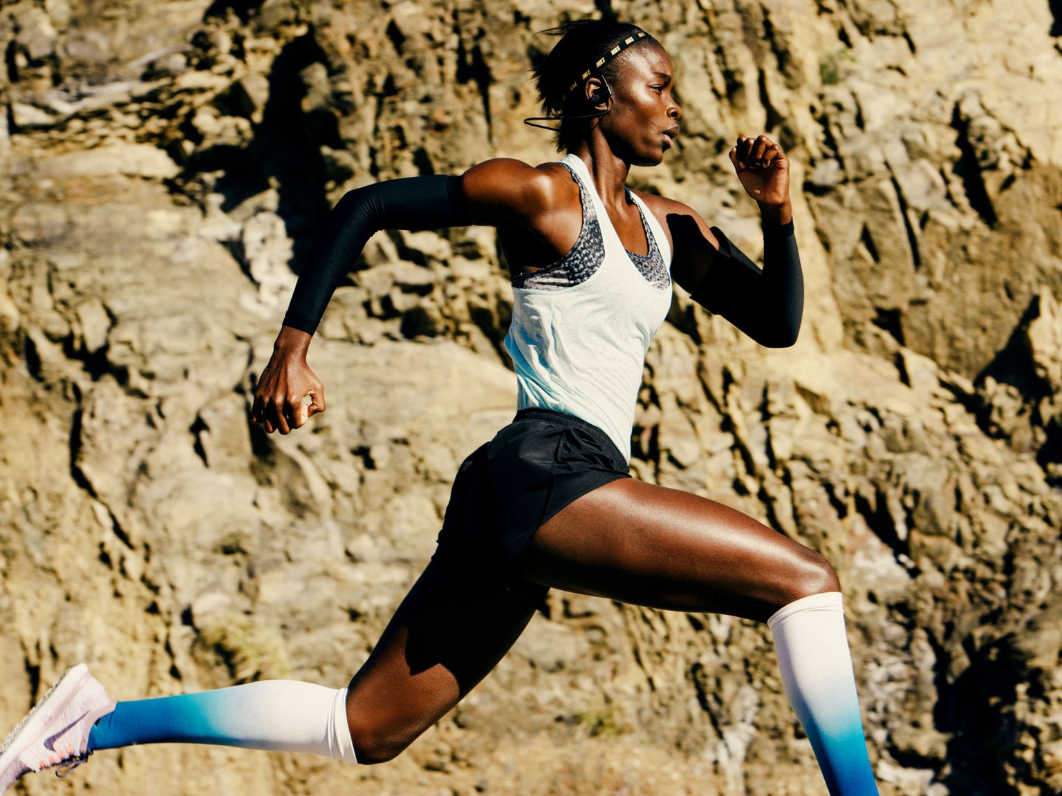 Athletic woman running outdoors. Dark-skinned female runner strides against a rocky landscape, wearing athletic wear and compression socks.