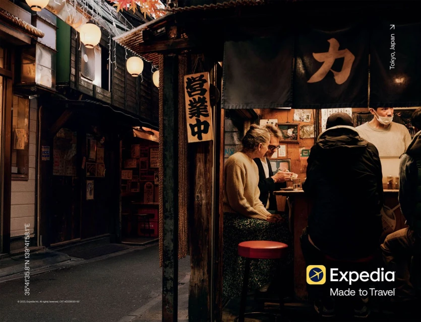 Image shows a Tokyo street scene with people at a small restaurant, illuminated by lanterns, creating an inviting and warm atmosphere.