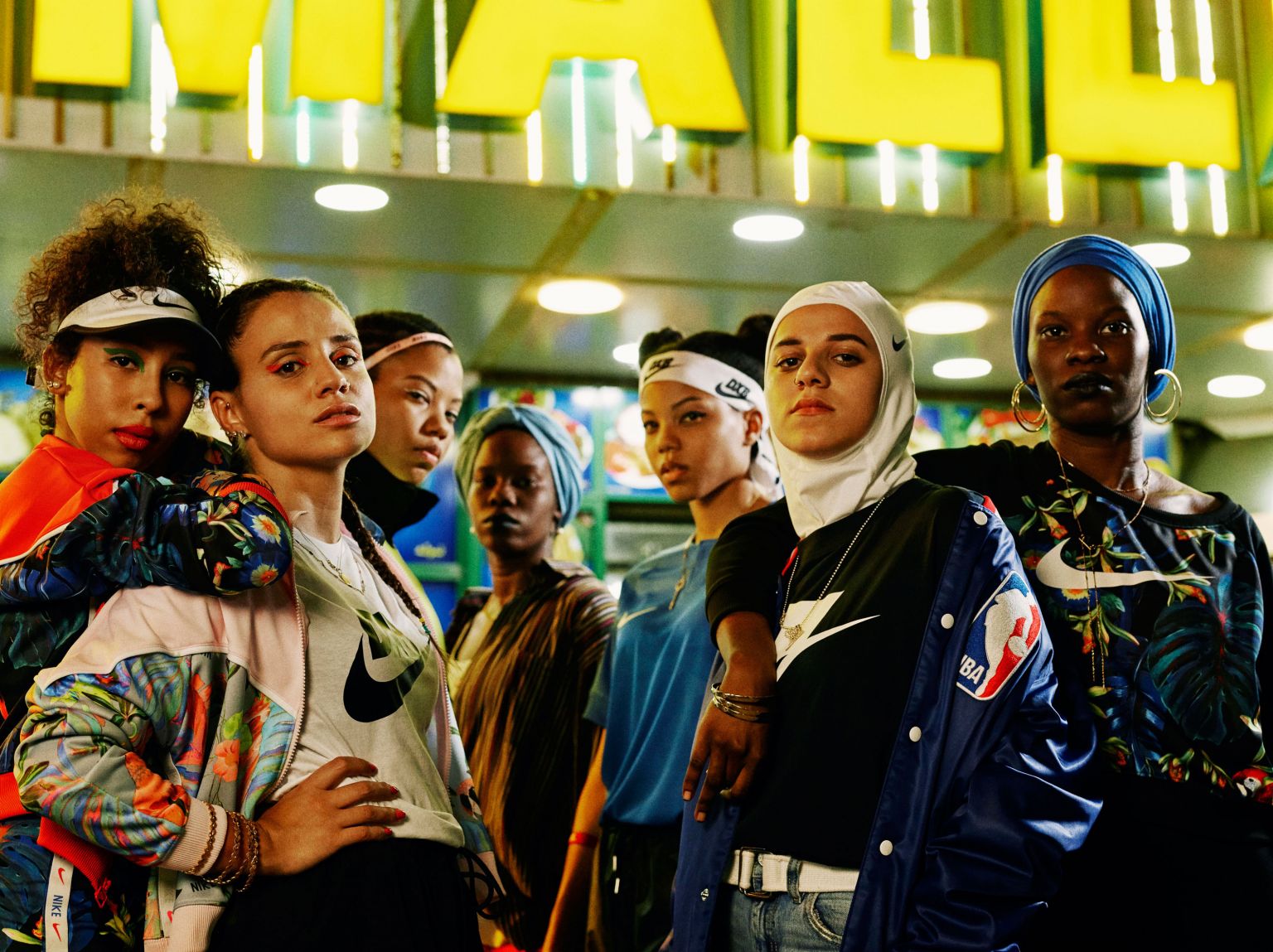 Group of diverse women in fashionable sportswear including tops, jackets, and head coverings pose under a bright sign in an urban environment.