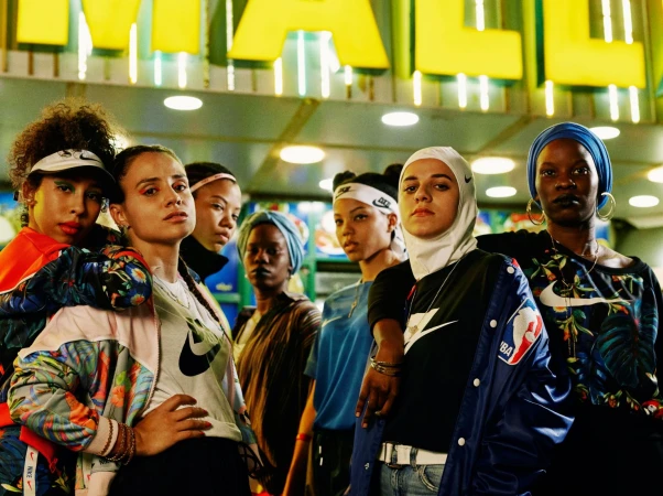 Group of diverse women in fashionable sportswear including tops, jackets, and head coverings pose under a bright sign in an urban environment.
