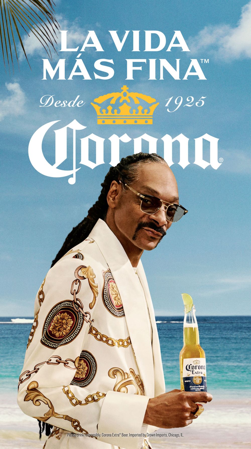 Snoop Dogg stands on a beach, wearing a patterned suit and sunglasses, holding a bottle of Corona beer with a lime.