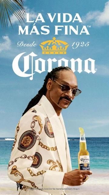 Snoop Dogg stands on a beach, wearing a patterned suit and sunglasses, holding a bottle of Corona beer with a lime.