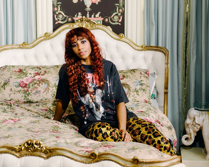 Stylish woman with auburn hair poses on a floral bed in a black band tee and leopard-print pants near elephant statue.