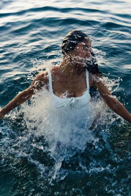 Woman in a white dress submerged in the ocean, captured in a medium shot with water splashing around her. Clear blue water surrounds her.