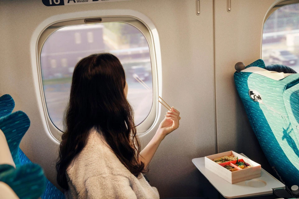 Brunette woman enjoys a bento box on a train, gazing out the window as scenery passes by, capturing a moment of travel and leisure.