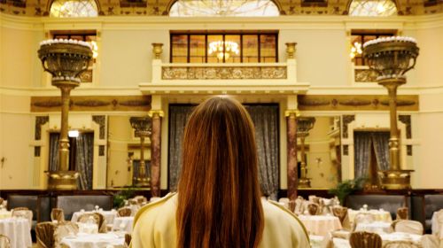 Back view of a woman with long brown hair standing in an ornate ballroom with set tables and chandeliers, facing away from the camera.