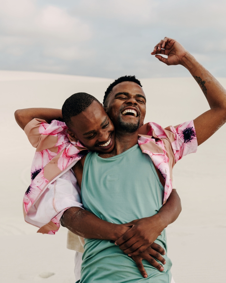 Two Black men joyfully embrace on sand dune, dressed in a floral shirt and tank top, with a bright, sunny atmosphere.