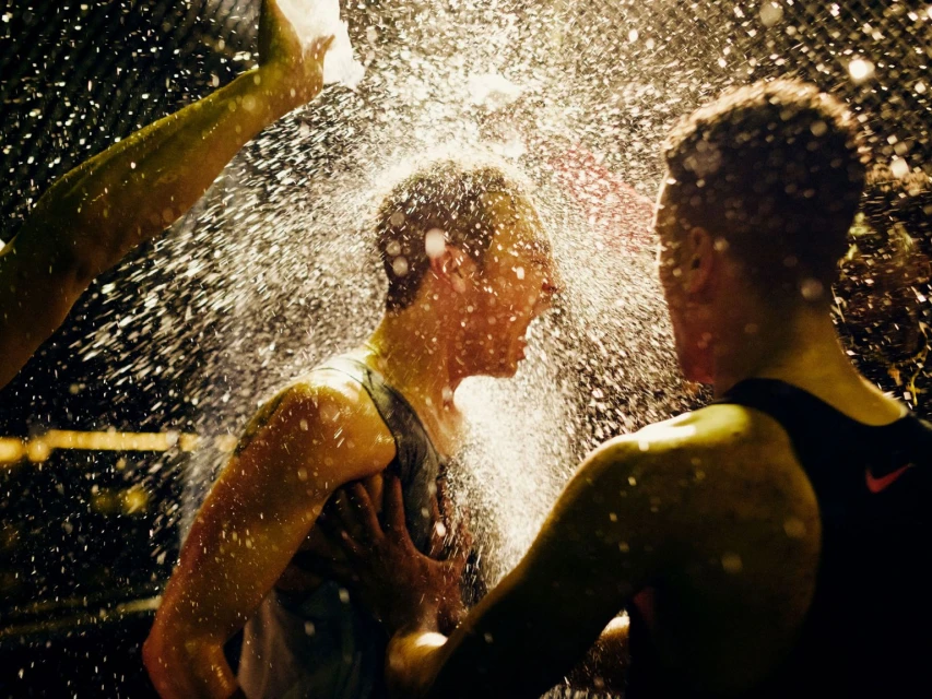 Two athletes celebrate a victory outdoors, doused in water. The individuals wear athletic gear and are illuminated by backlighting.