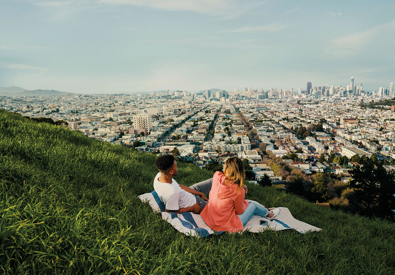 A couple sits on a blanket on a grassy hill overlooking San Francisco. The city is visible in the distance on a clear, sunny day.