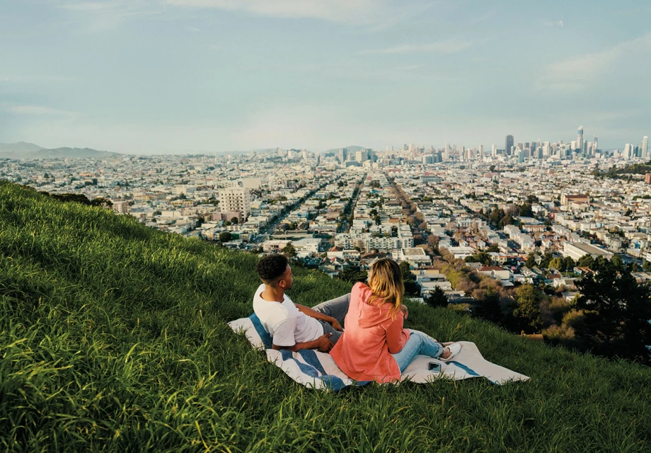 A couple sits on a blanket on a grassy hill overlooking San Francisco. The city is visible in the distance on a clear, sunny day.