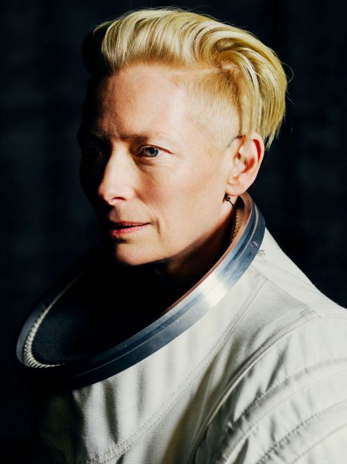 Close-up of an adult female with short, blonde, slicked-back hair, wearing a white space suit with a silver collar in a studio.
