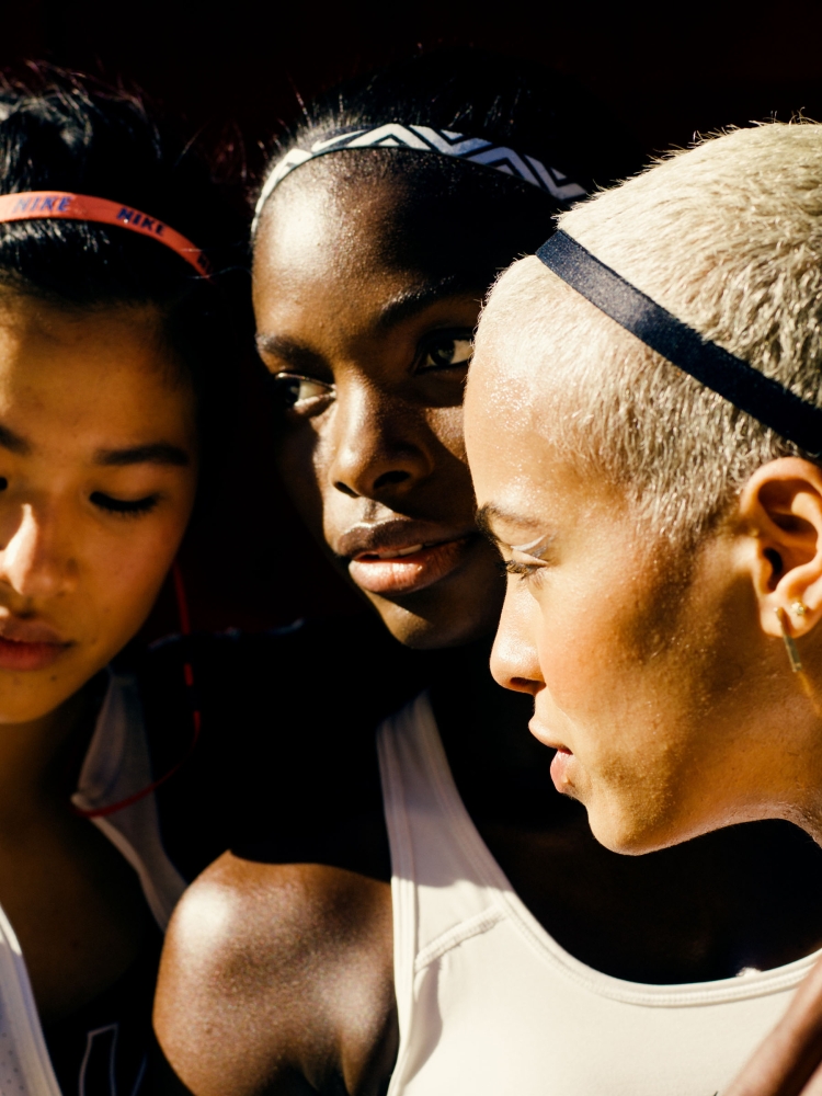 Close-up of three diverse young female athletes wearing athletic wear and headbands against a dark background, lit with soft light.