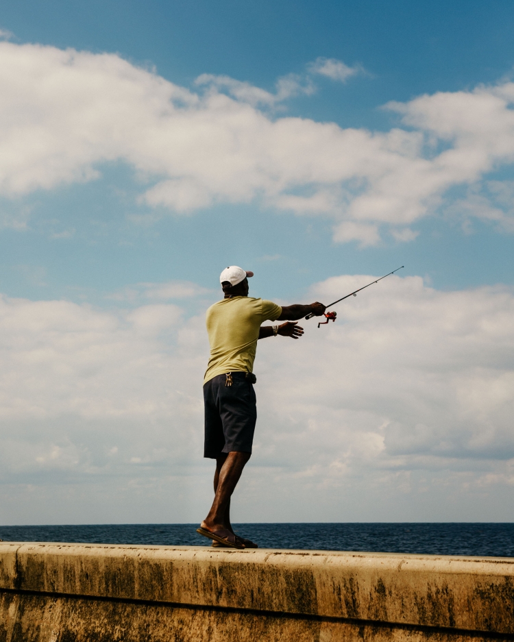 Adult man fishing with a rod and reel off a sea wall with ocean and sky in the background on a sunny day, wearing a hat.