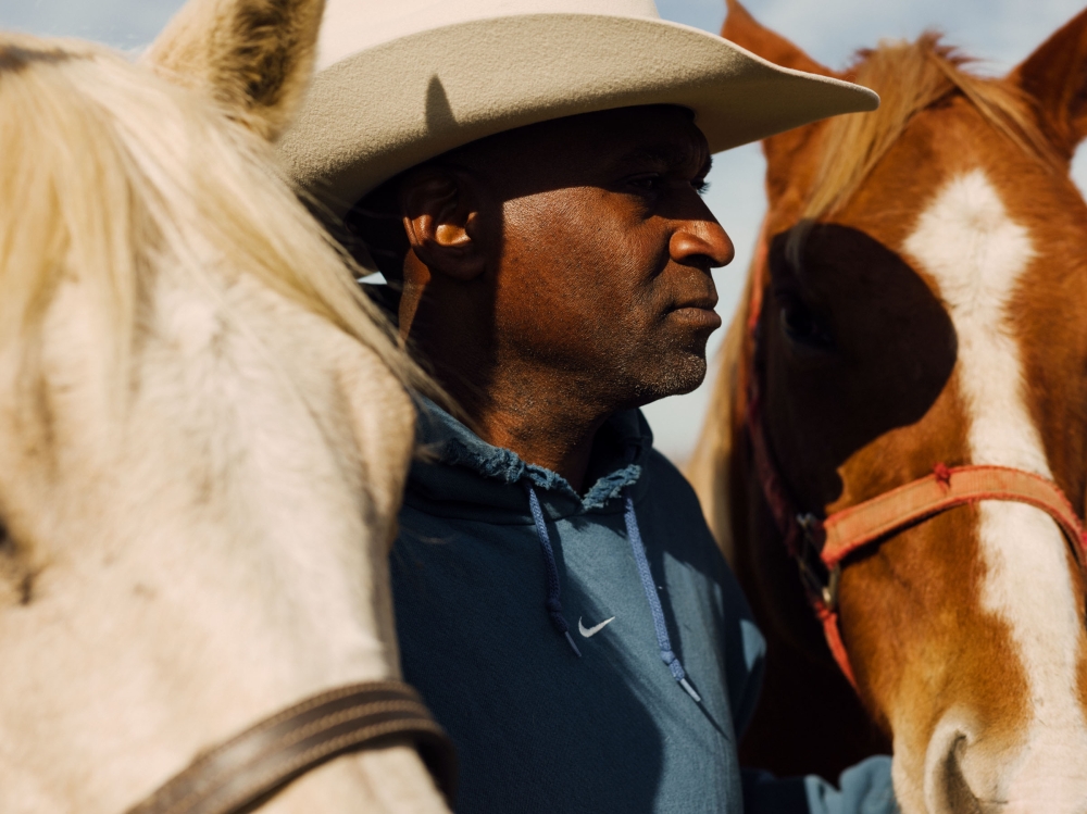 Close-up of a man in a cowboy hat with two horses, one cream-colored and one brown, in an outdoor setting with natural light.