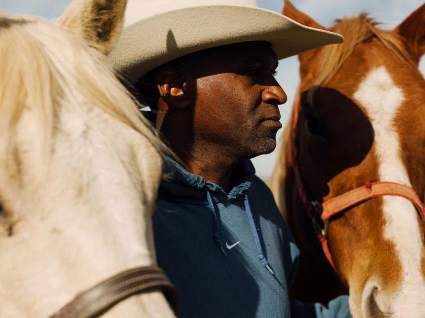 Close-up of a man in a cowboy hat with two horses, one cream-colored and one brown, in an outdoor setting with natural light.