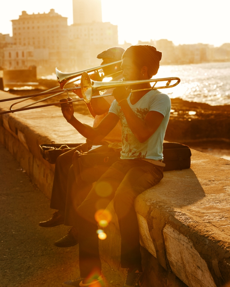 Two musicians play trombones on a concrete ledge by the sea, bathed in the warm glow of the setting sun with buildings in the background.