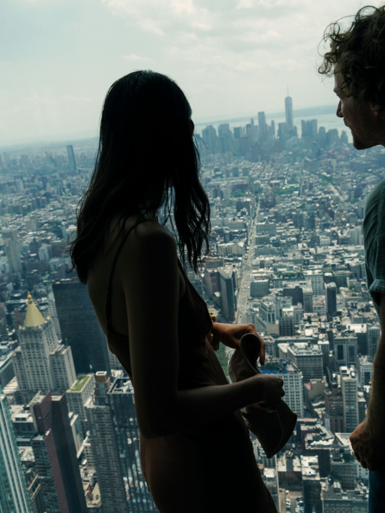 Silhouetted woman and man stand overlooking New York City skyline from a high vantage point on a sunny day, creating a striking urban view.