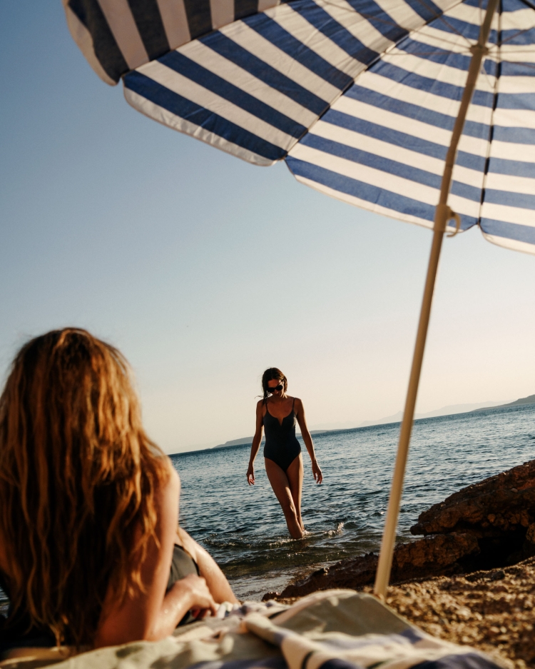 Two women at the beach: one in the water in a swimsuit, the other lounging under a blue and white striped umbrella on the sand.