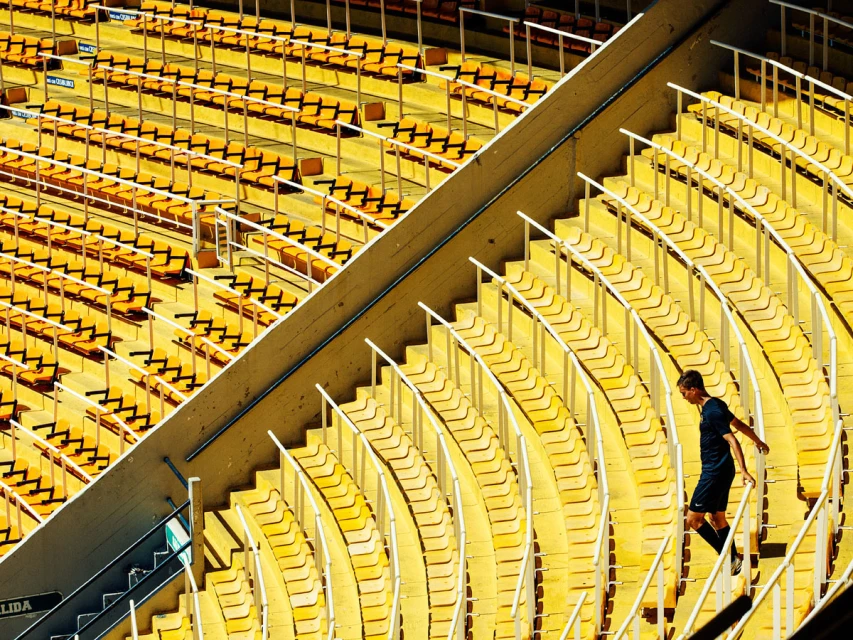 Stadium seating with a lone adult male walking down the stairs on a sunny day, bright yellow seats, and white railings.