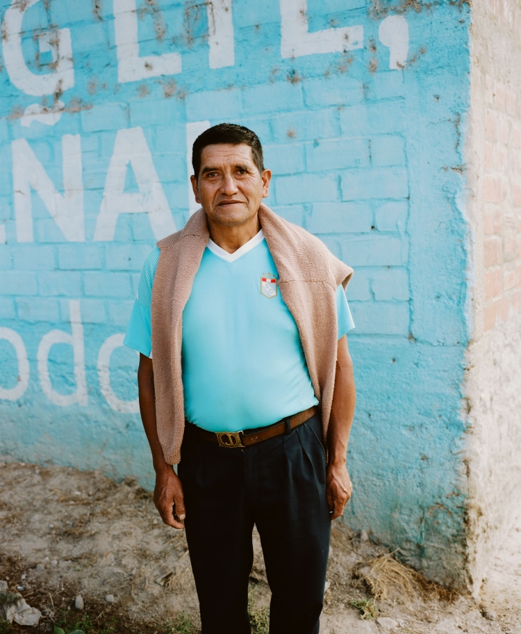 Portrait of a middle-aged man in a blue shirt and dark pants, wearing a tan sweater, standing against a light blue brick wall with white paint.