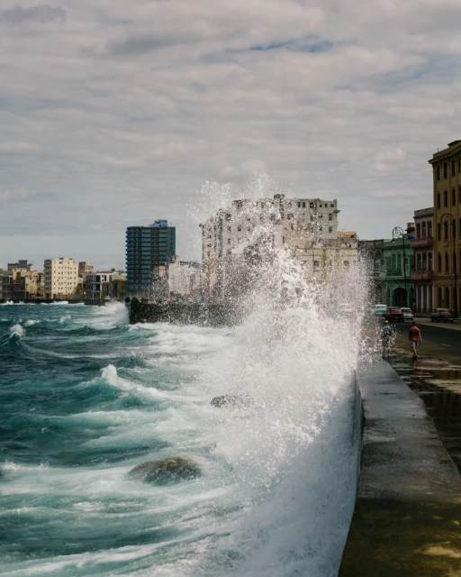 Waves crashing on the Malecón seawall in Havana, Cuba, with city buildings, a cloudy sky, and a pedestrian in the background.