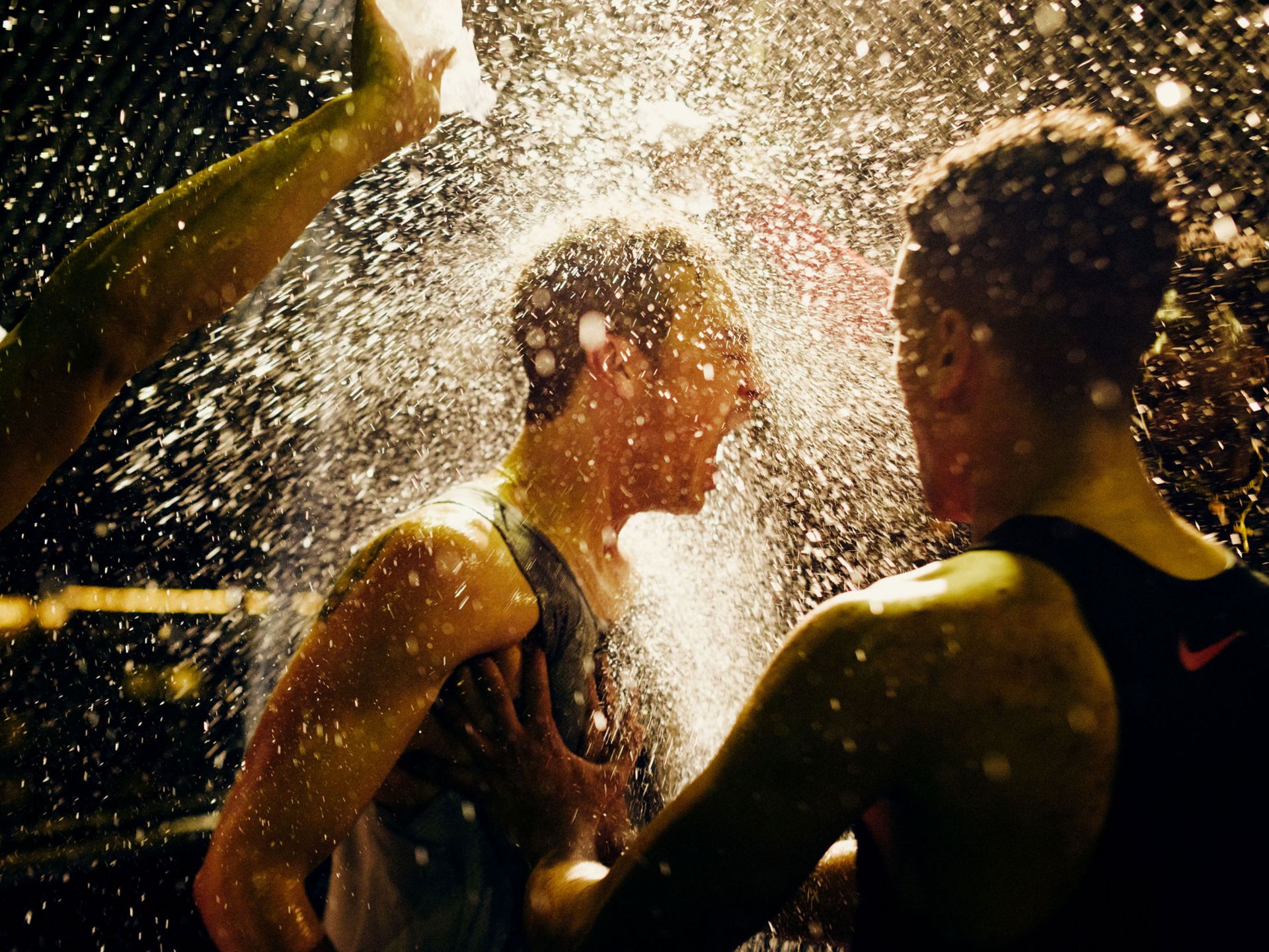 Two adult male athletes drenched in water, one with eyes closed and hands at his chest, possibly celebrating a victory in dim light.