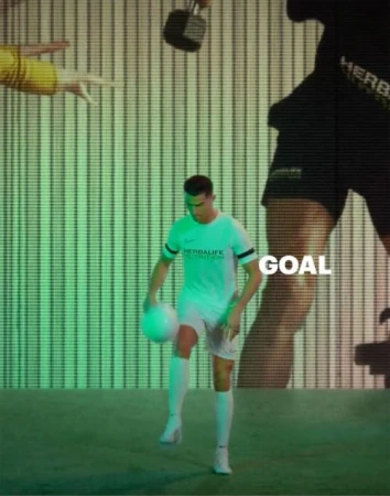 Male soccer player in Herbalife uniform juggling a soccer ball indoors on a green field with a digital display background.