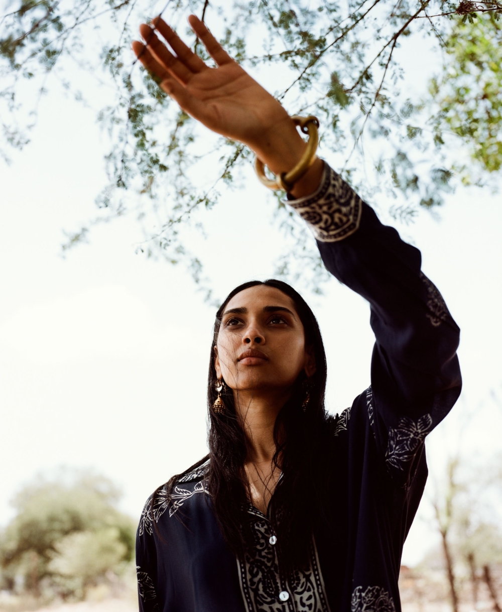 Adult woman in patterned shirt reaches toward tree leaves in outdoor setting with bright sky. Model wears bangle and earrings.