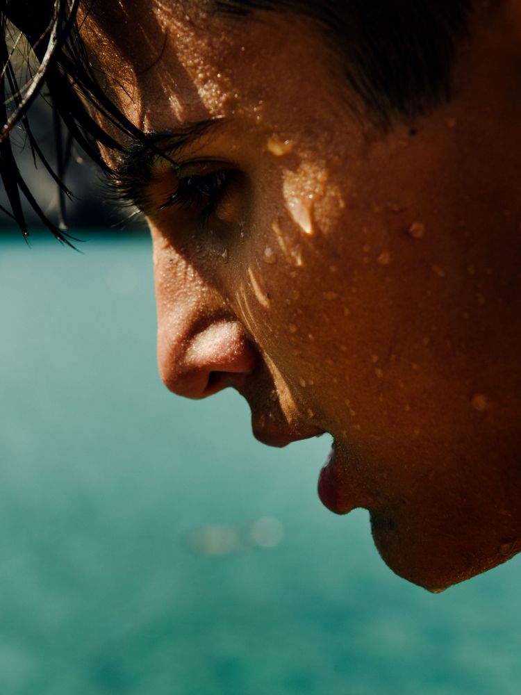 Profile of a wet man's face against a blurred turquoise backdrop, emphasizing skin texture and water droplets in a close-up shot.