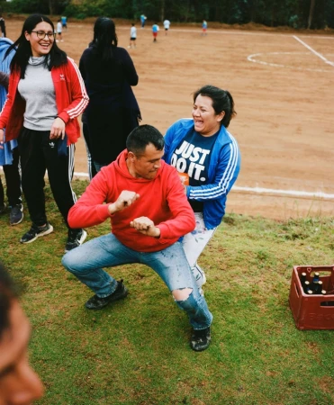 Group of adults laughing and interacting outdoors in a grassy area. Man in a red hooded sweatshirt is crouched. Woman in a blue top is behind him.