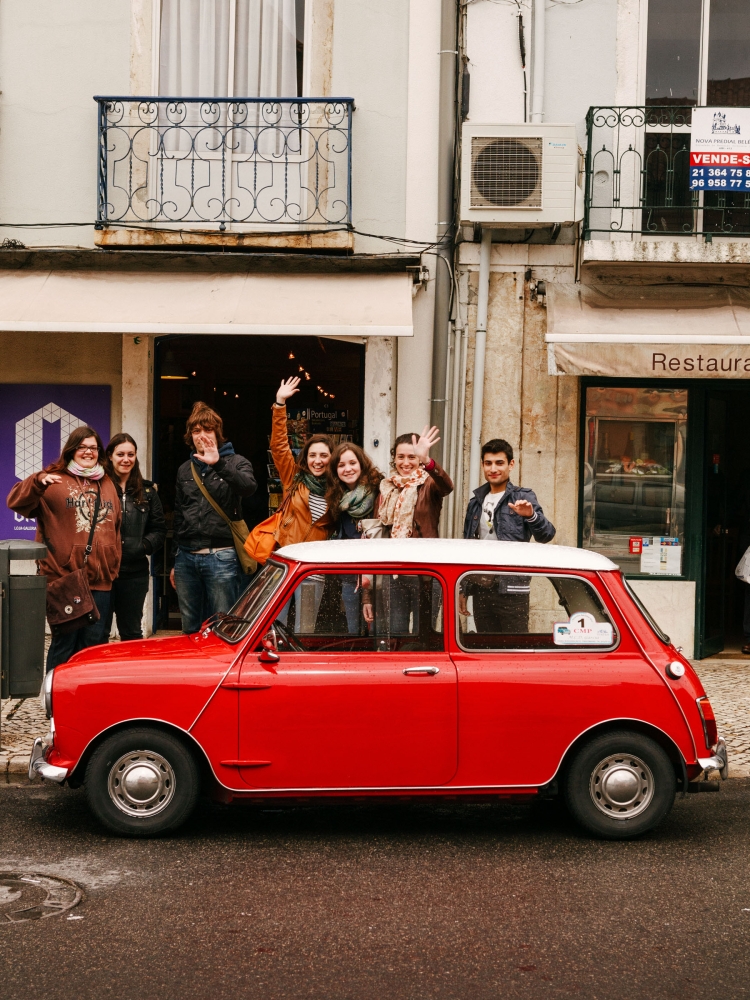 Group of six people posing with a red Mini Cooper on a city street in front of a building with an iron balcony and awnings.