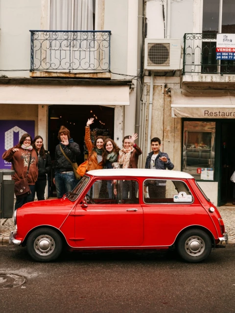 Group of six people posing with a red Mini Cooper on a city street in front of a building with an iron balcony and awnings.