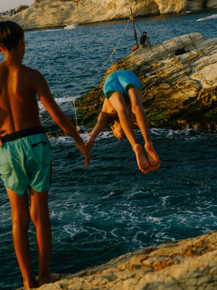 Two boys diving into the blue sea from rocky shore. One boy is upside down, diving, while the other watches, with a rocky cliff backdrop.