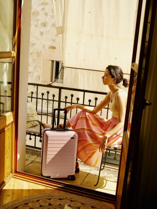 Woman sitting on a balcony next to a pink suitcase with a stone wall background