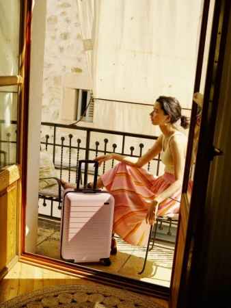 Woman sitting on a balcony next to a pink suitcase with a stone wall background