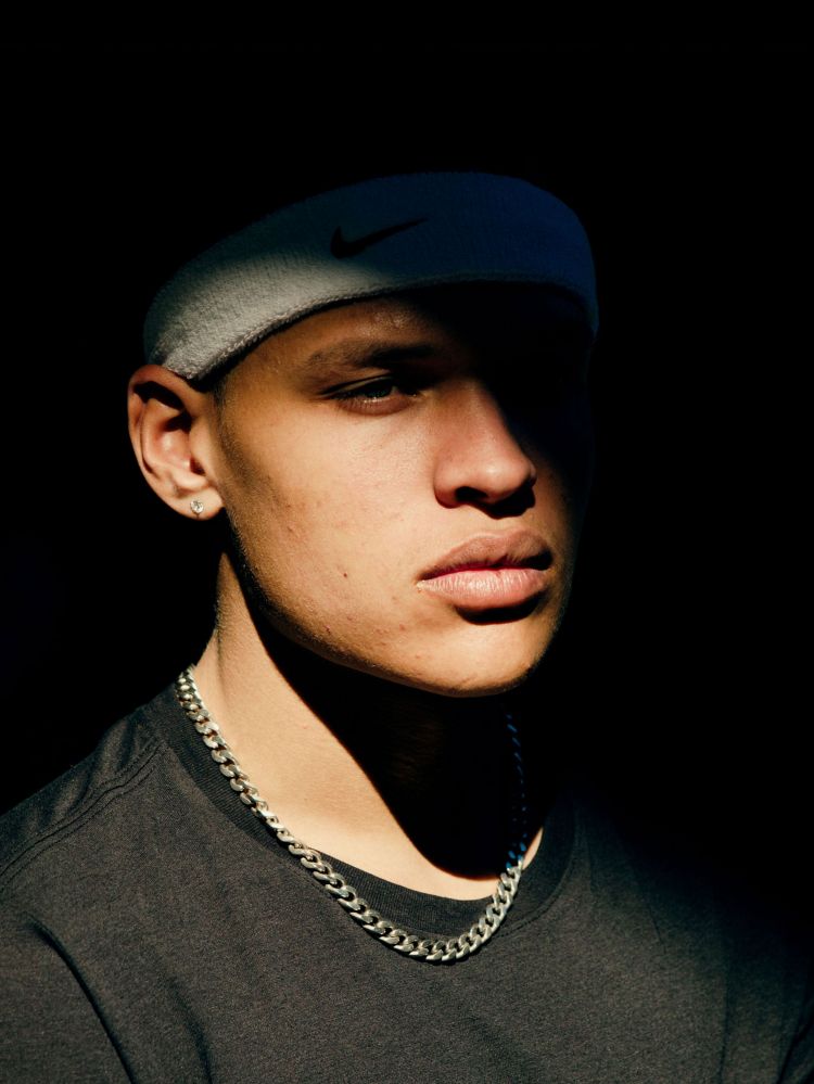 Close-up portrait of a young adult male with a gray headband, silver chain necklace, and a dark t-shirt, set against a dark background.
