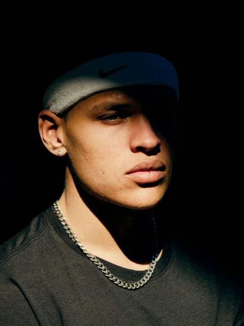 Close-up portrait of a young adult male with a gray headband, silver chain necklace, and a dark t-shirt, set against a dark background.