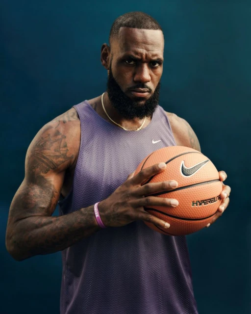 Basketball player LeBron James holds a basketball while wearing a purple mesh tank top in a medium studio shot on a blue background.
