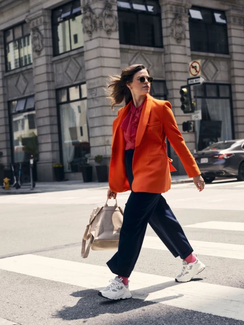 Woman in orange blazer walks across crosswalk in city. Holding tote bag, sunglasses, pink socks, white sneakers. Architecture background.