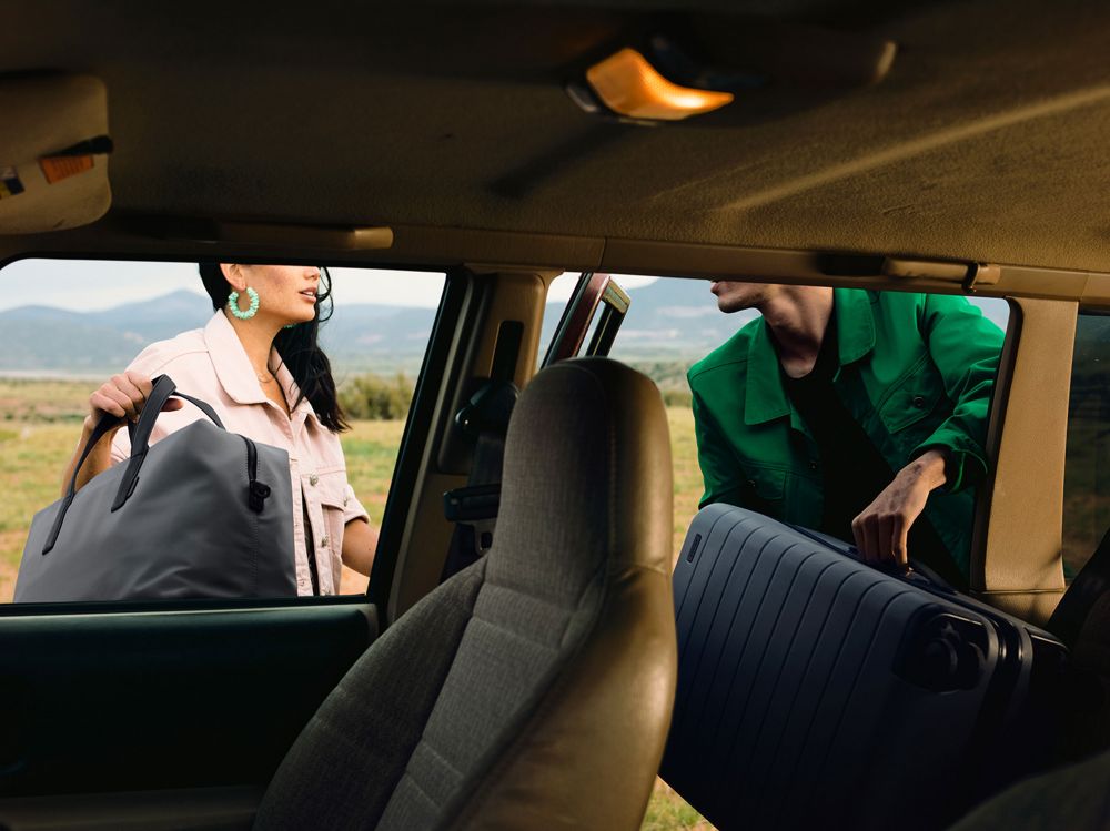 Two people loading luggage into the back of a car in a rural setting with green grass and mountains in the background, travel concept.