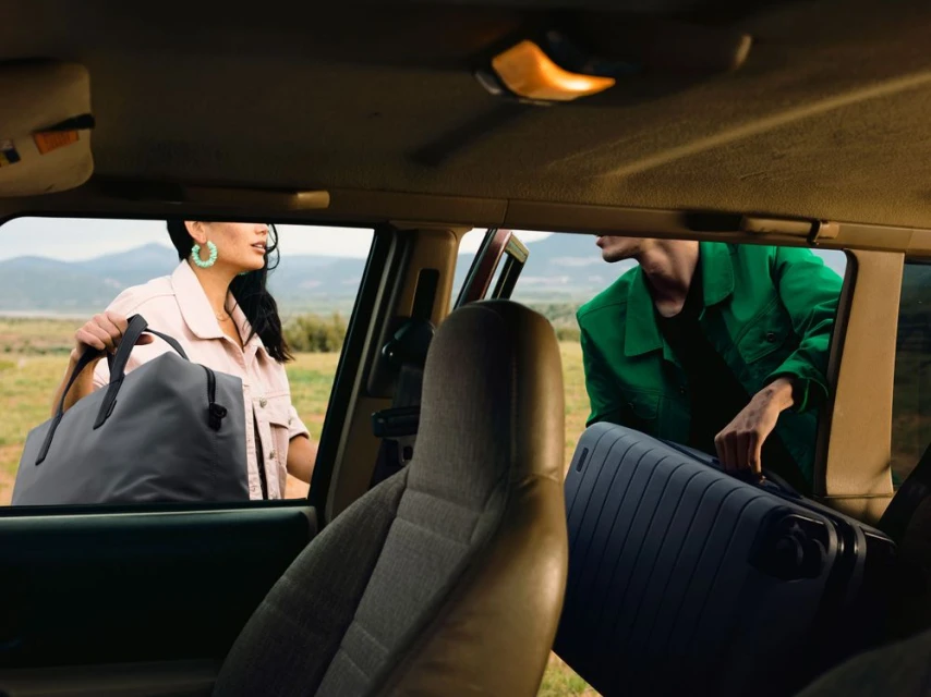 Two people loading luggage into the back of a car in a rural setting with green grass and mountains in the background, travel concept.