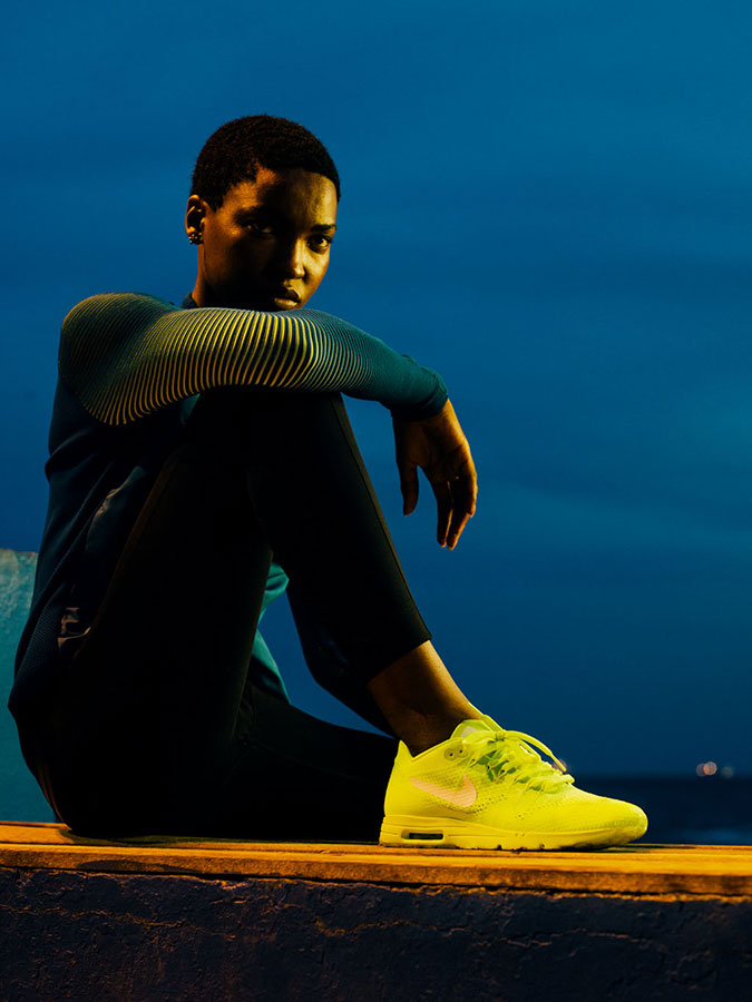 An athletic woman in sportswear is sitting in a neon-lit outdoor environment wearing vibrant neon yellow Nike sneakers.