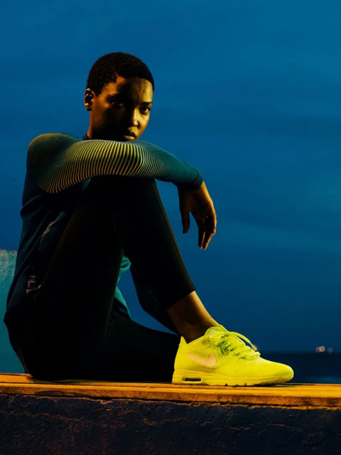 An athletic woman in sportswear is sitting in a neon-lit outdoor environment wearing vibrant neon yellow Nike sneakers.