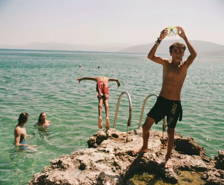 A group of young people are swimming in the ocean on a sunny day, one diving off rocks, another holding swimming goggles above head.