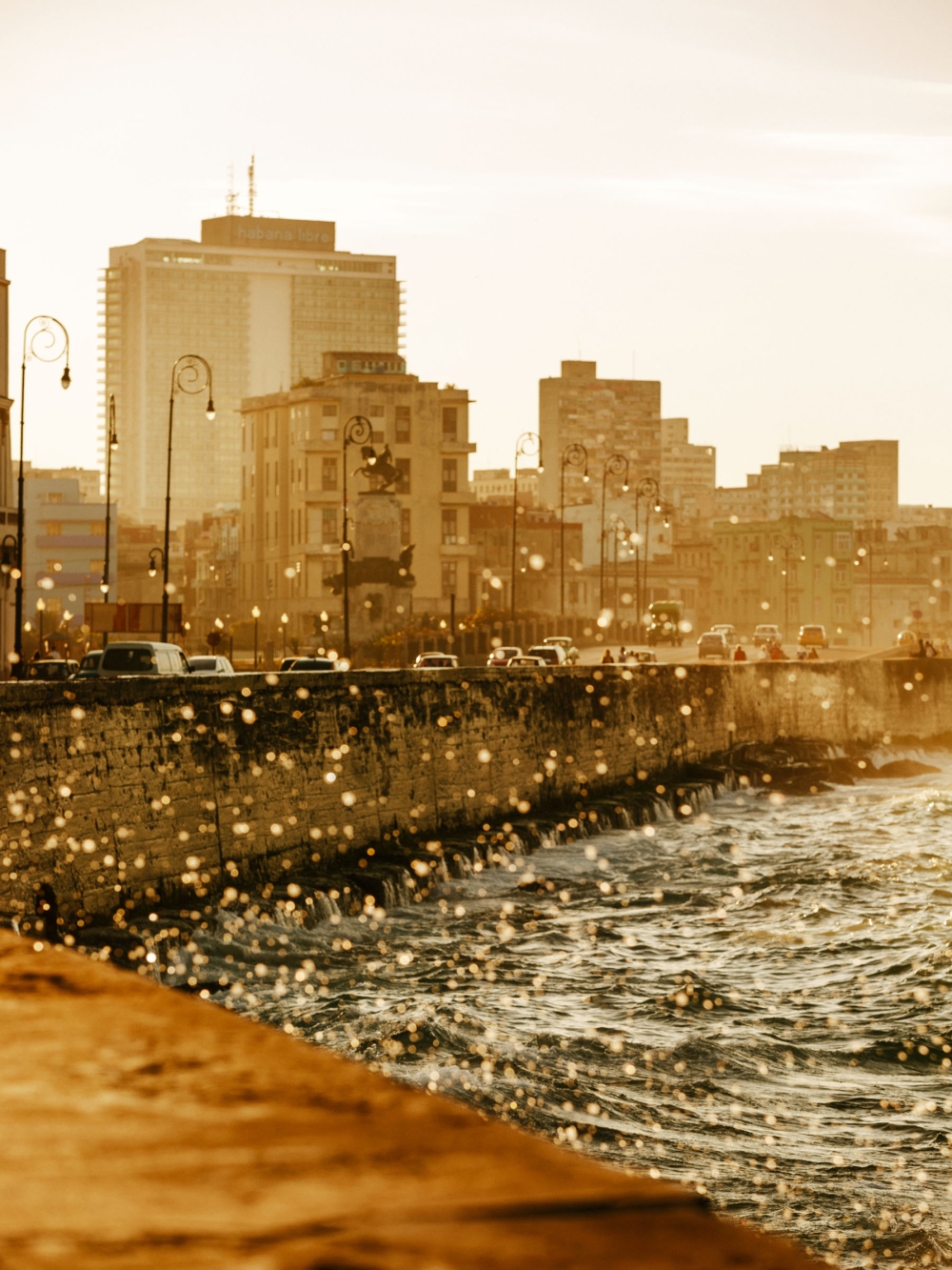 Cityscape view of Havana, Cuba featuring vintage cars driving along the Malecon seawall with aged buildings under golden light.