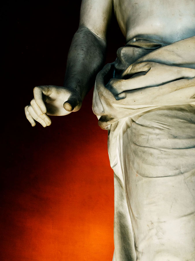 Close-up of a marble sculpture featuring a draped figure with visible hand and arm details, set against a dark red and black background.
