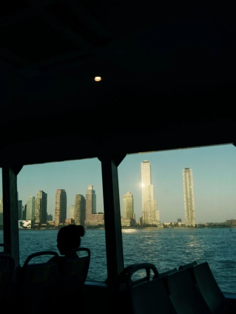 City skyline view from a ferry with a silhouetted figure looking out the window at buildings and water on a sunny day.