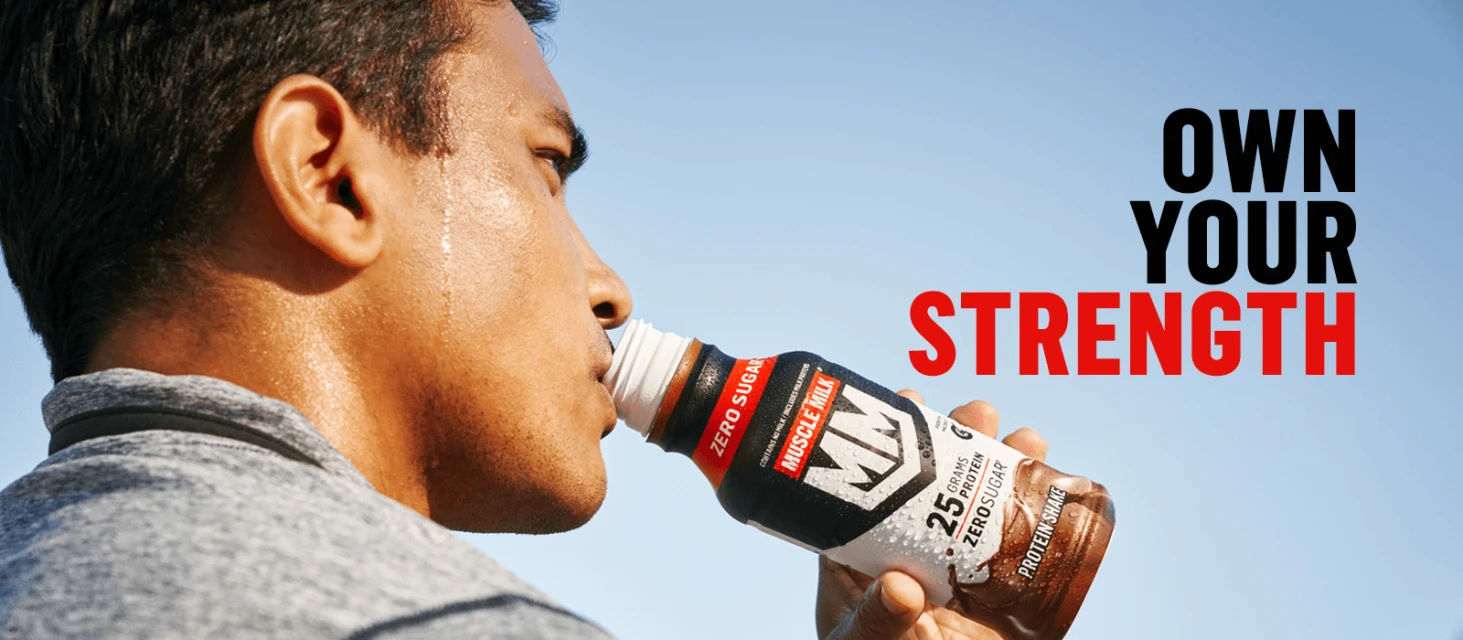 Adult male drinking Muscle Milk protein shake outdoors against blue sky background, showcasing fitness and nutrition for an active lifestyle.