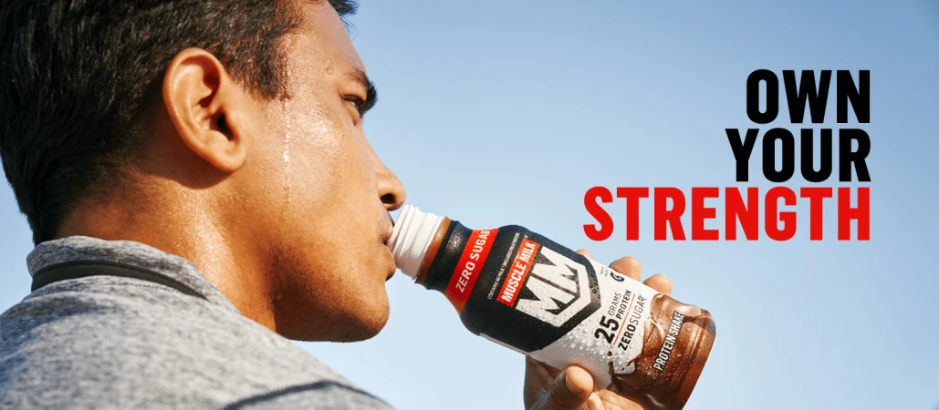 Adult male drinking Muscle Milk protein shake outdoors against blue sky background, showcasing fitness and nutrition for an active lifestyle.